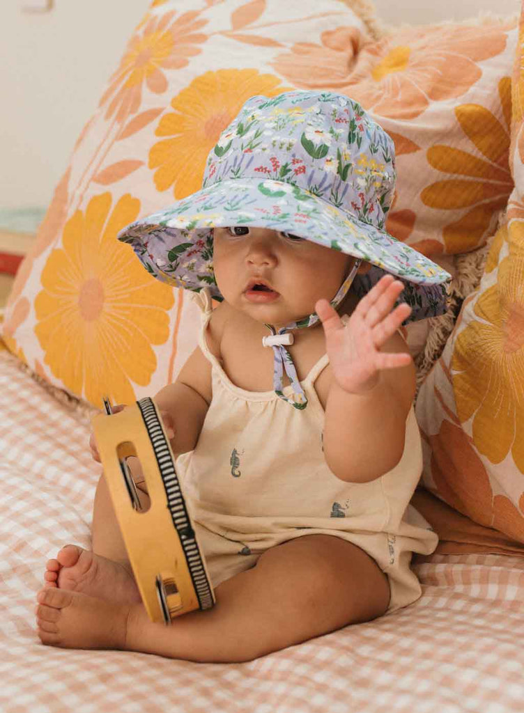 Baby wearing a floral sun hat sitting on a checkered blanket with a toy.