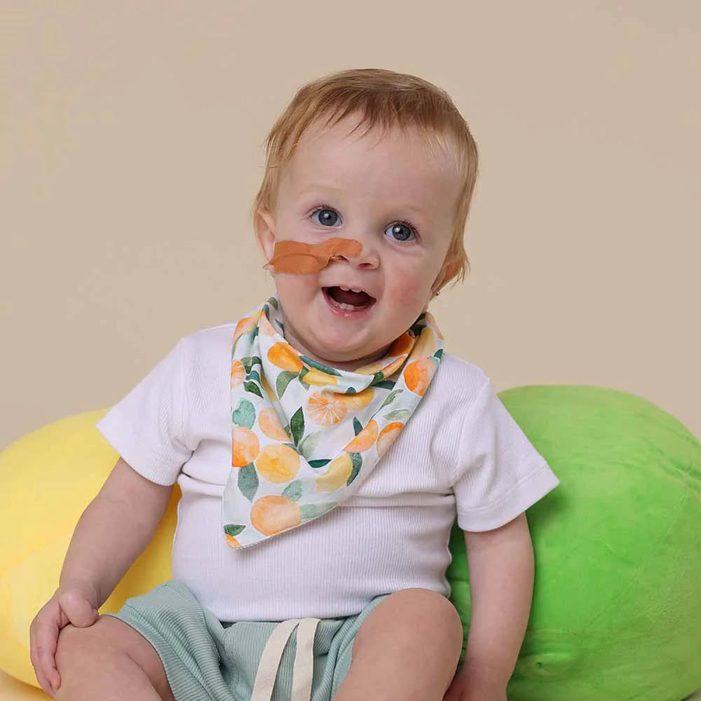 Baby sitting on a colorful chair wearing a lemon patterned bib with a neutral background